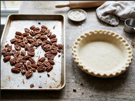 Toasted pecan halves cooling on a sheet pan next to an unbaked pie crust in a 9-inch plate