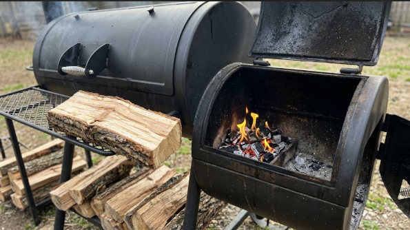 Offset smoker with the firebox open, a small clean fire burning, and a split of oak ready to add