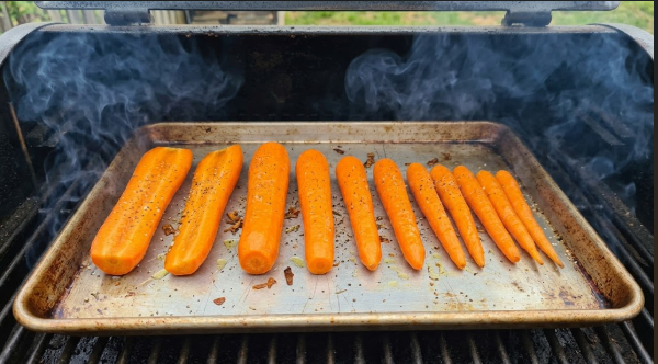 Seasoned carrots in a single layer on a sheet pan inside the pellet grill with thin blue-gray smoke rising
