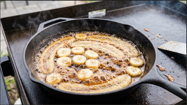 Banana slices cut-side down in bubbling brown sugar sauce in a cast iron skillet on the Blackstone