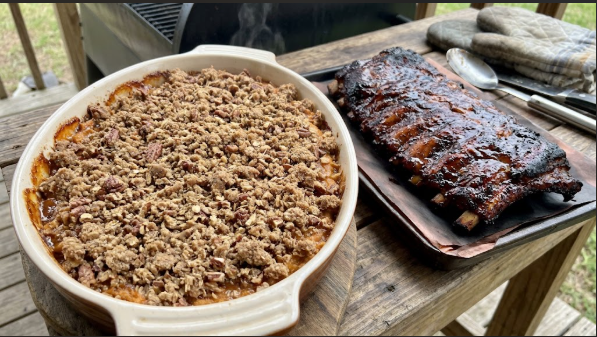 Finished sweet potato casserole in the pan next to smoked ribs, topping golden and bubbling