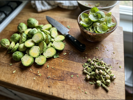 Halved sprouts on a cutting board next to a bowl of loose leaves and a small pile of trimmed stems