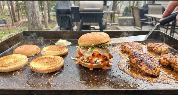 Buffalo chicken sandwich on a griddle with toasted buns and chicken sizzling nearby
