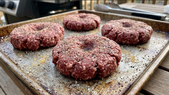 Raw burger patties with a thumb dimple pressed into the center, seasoned and ready for the grill