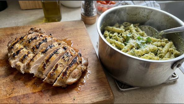 Sliced grilled chicken thighs with visible grill marks, resting on a cutting board next to a pot of pesto pasta