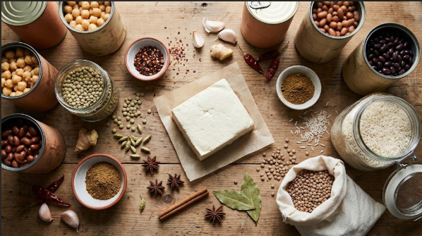 Pantry shelf or counter flat-lay of canned beans, lentils, rice, spices, and a block of tofu