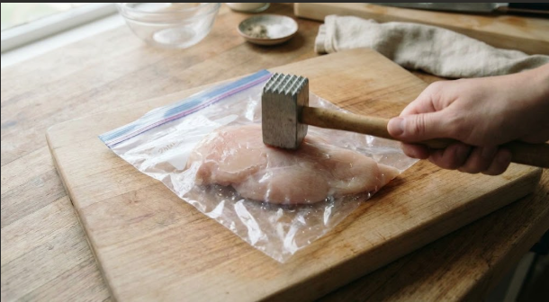 A chicken breast inside a zip-top bag being pounded with a mallet, with the thick end already flattened