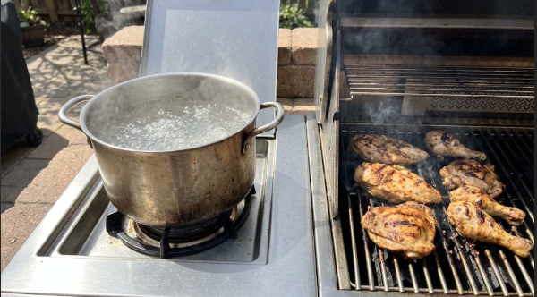 A pot of salted boiling water on a side burner next to a grill with chicken cooking