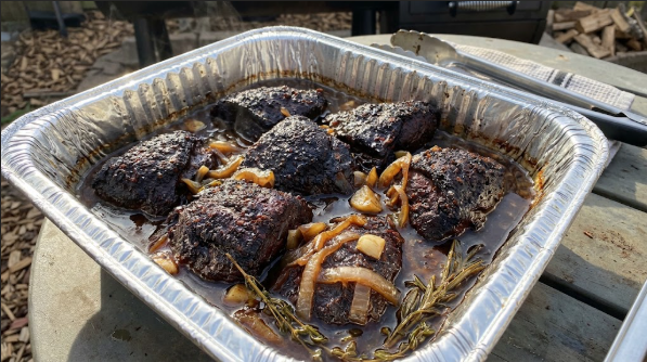 Bark-covered beef cheeks nestled in a foil pan with onions and braising liquid