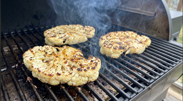 Cauliflower steaks on a pellet grill grate with visible browning and light smoke