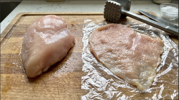 Raw chicken breasts on a cutting board, one pounded flat next to one untouched, to show the thickness difference