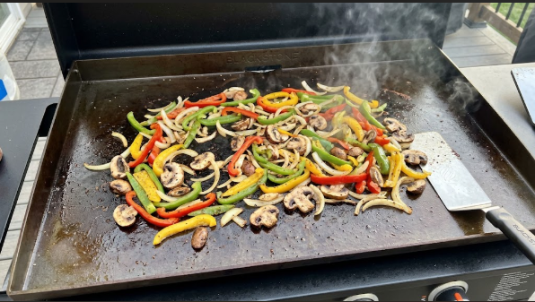 Sliced peppers, onions, and mushrooms sizzling in a single layer on a Blackstone griddle