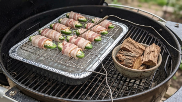 Grill setup with a wire rack over a foil pan holding bacon-wrapped poppers, a thermometer probe, and a bowl of pecan wood chunks