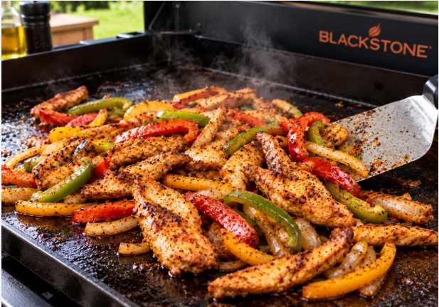 Chicken strips and sliced peppers sizzling on a Blackstone griddle with a metal spatula