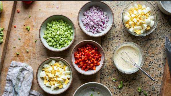 Diced celery, red onion, bell pepper, and chopped eggs in small bowls next to a whisked dressing, shot from above