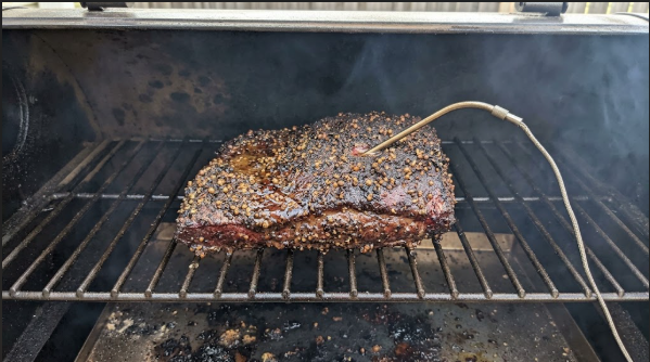 Corned beef flat on the smoker grate with a dark pepper bark and a probe in place
