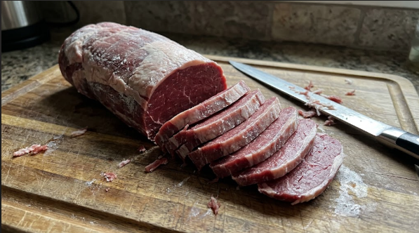 Partially frozen eye of round roast on a cutting board with a long slicing knife and even ¼-inch strips fanned out