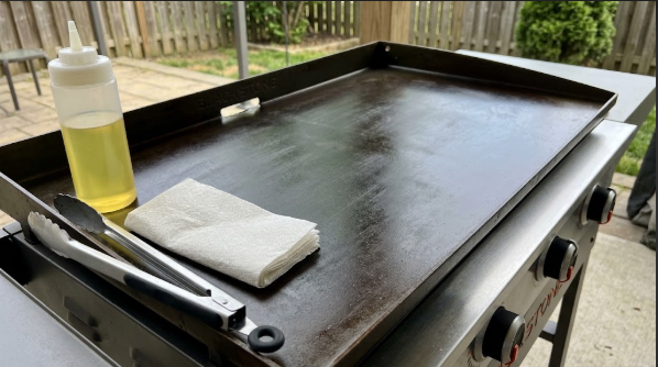 Squeeze bottle of canola oil next to a clean Blackstone griddle top with tongs and folded paper towels