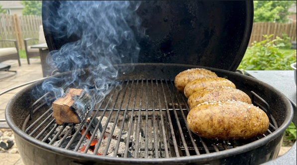 Four russets on the indirect side of a grill with the lid open and a chunk of wood smoking nearby