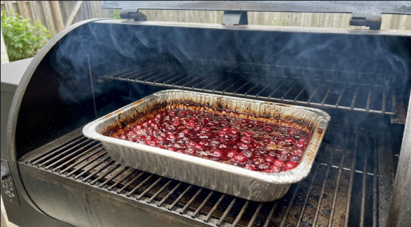 Foil pan of cranberry sauce on the smoker grate with thin blue smoke